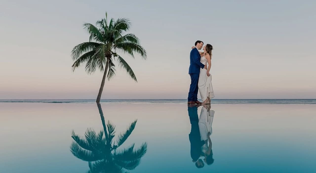 Bride and groom facing the sea, reflected in the infinity pool at sunset